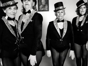 Four older women wearing costumes of sequined tuxedo jackets, tophats and tights stare at the camera