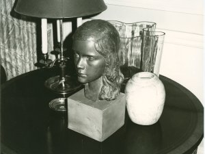 Black-and-white photo shows a table topped with a statue of a girl's head, vase and lamp. 