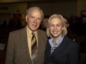 An older man in a tan suit coat and yellow shirt with a striped tied poses for a photo with a blond woman in a blue collared shirt and dark coat.