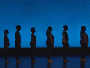 The silhouettes of eight people standing in profile on a stage can be seen against a dark blue backgroundst a 