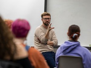 A man with a beard teaches at the front of an LGBTQ Studies class while wearing a tan sweater and glasses. A blank projector screen is lowered on the wall behind him.