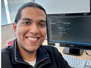 Jorge Gomez smiles in front of a computer monitor displaying programming text.