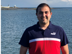 Bradey Liverio stands in front of body of water, wearing a navy, white, and red collared shirt