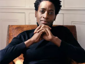 A seated woman with short black hair wears a black turtleneck and stares into the camera, her hands crossed beneath her chin.