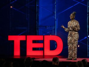 Kyra Gaunt stands on a darkly lit stage next to red letters that read TED