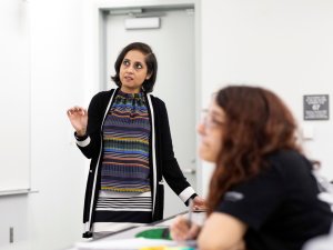 Sreya Kolay stands in front of a classroom teaching. A student sits out of focus in the foreground with long auburn hair and glasses.