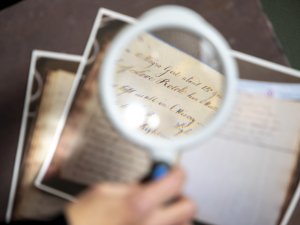 A magnifying glass is held up to a photocopy of an old poem written in elegant cursive script on a page that has been yellowed and faded from age.