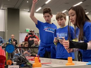 Two boys and a girl, all in blue tee shirts, stare down on robotics game board. 
