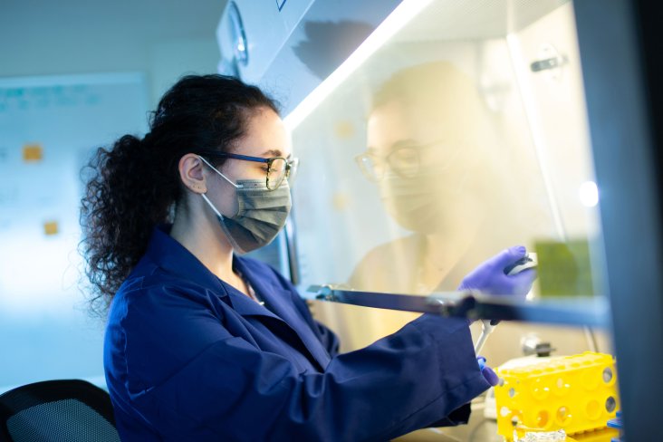 A researcher in a gray surgical mask, purple nitrile gloves and a navy blue lab coat works with samples inside a biology lab.