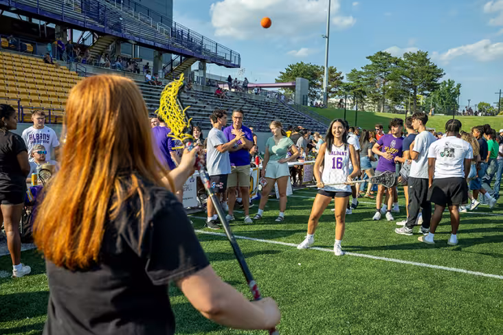 Two students play lacrosse with each other at Great DaneTopia while other attendees socialize and watch.