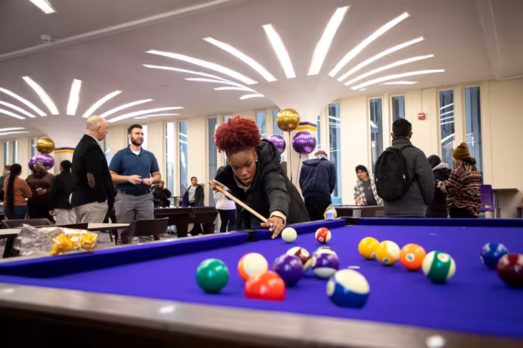 A student plays pool in the Campus Center Game Room. Other students socialize in the background.