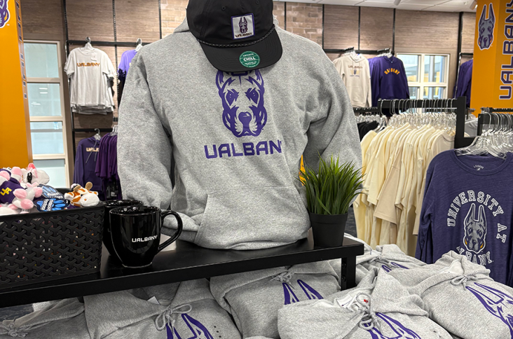 A mannequin wearing a gray UAlbany sweatshirt and a black UAlbany hat, sitting above stacks of gray UAlbany sweatshirts in Academien's Bookstore.