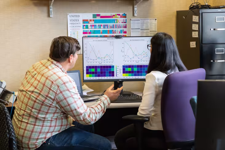 Two professionals sit in front of a computer monitor displaying data graphics. One individual gestures as he speaks.