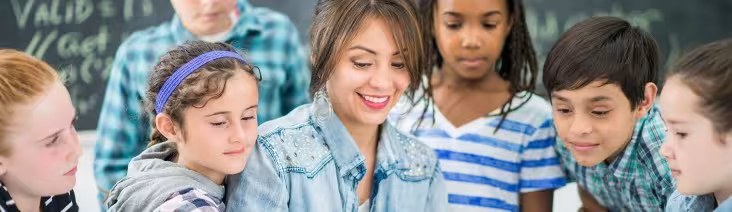 A teacher looking at a laptop while surrounded by a group of young students.