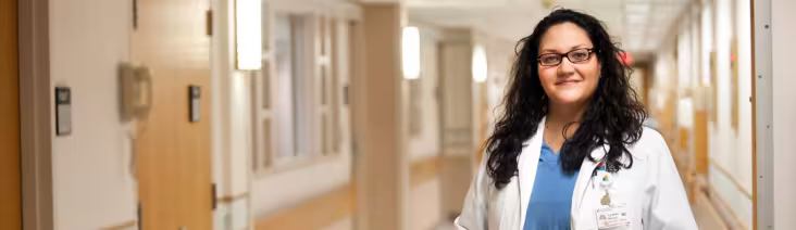 UAlbany alum Cynthia Bernal, MD standing in a hospital hallway wearing physician's scrubs and lab coat.