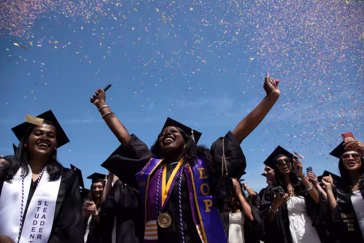 A student in Commencement regalia smiles widely and throws hands in the air in celebration.