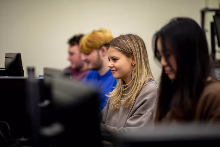 Four students sit working in a row of desktop computers. The camera is focused on one student who is smiling as she works.