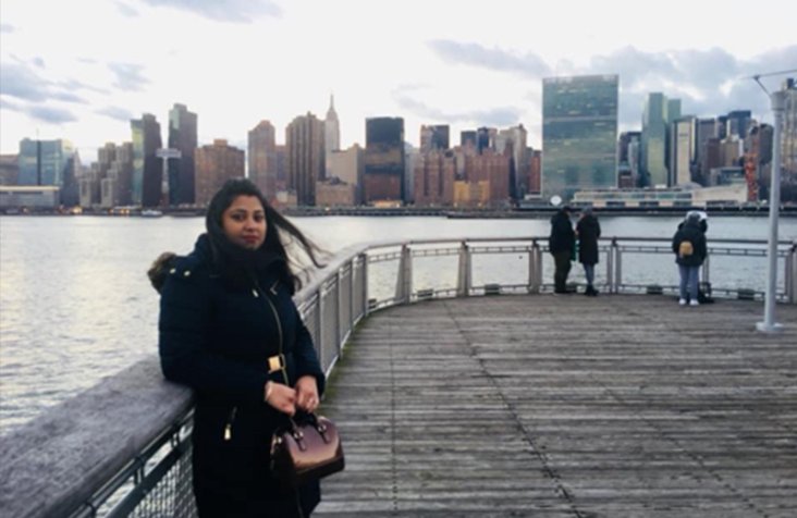 Farial Nur Maysha stands in a black coat on a wooden pier in front of a river and city skyline.