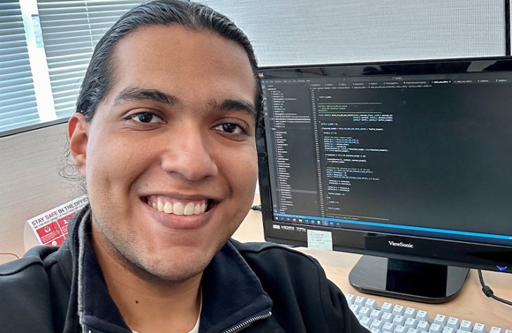 Jorge Gomez smiles in front of a computer monitor displaying programming text.