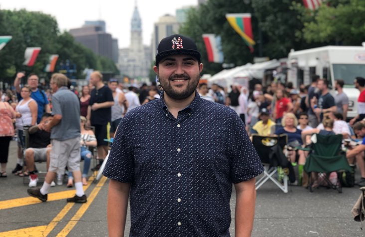 Bradey Liverio stands in a city street wearing a Yankees cap and a blue button-up short-sleeved shirt, surrounded by a crowd of people.