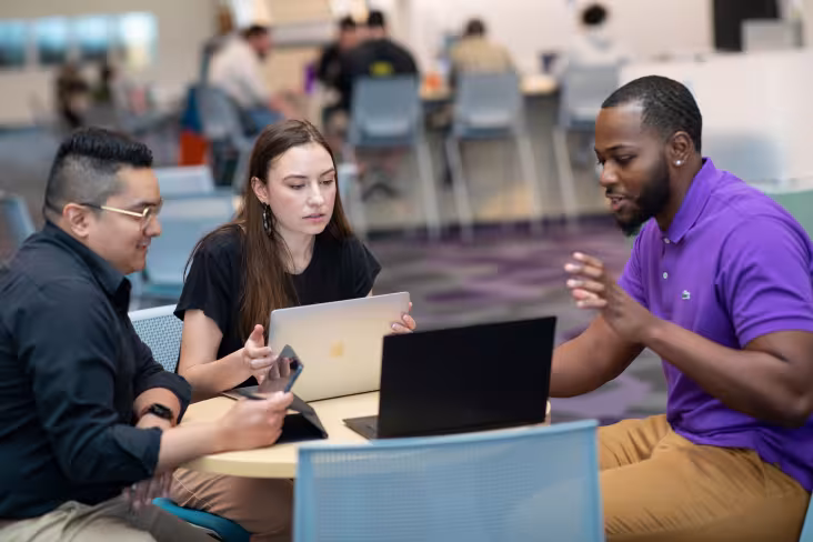 Three students talk and collaborate while sitting at a circular table with open laptops.