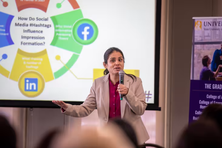 A graduate student holding a microphone presents a slide on social media to a seated crowd.