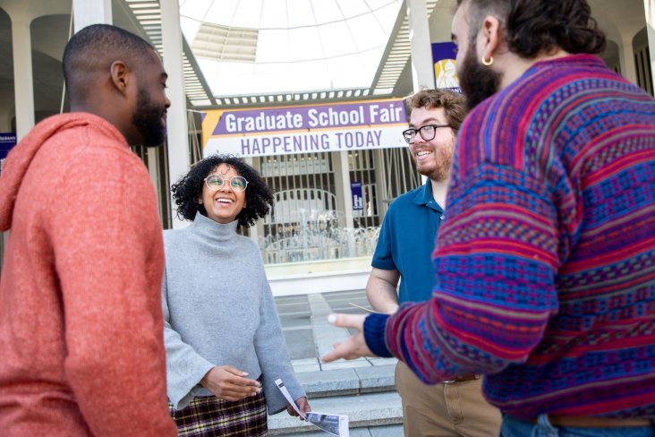 Four students smile and talk in front of a sign that says, "Graduate School Fair Happening Today."