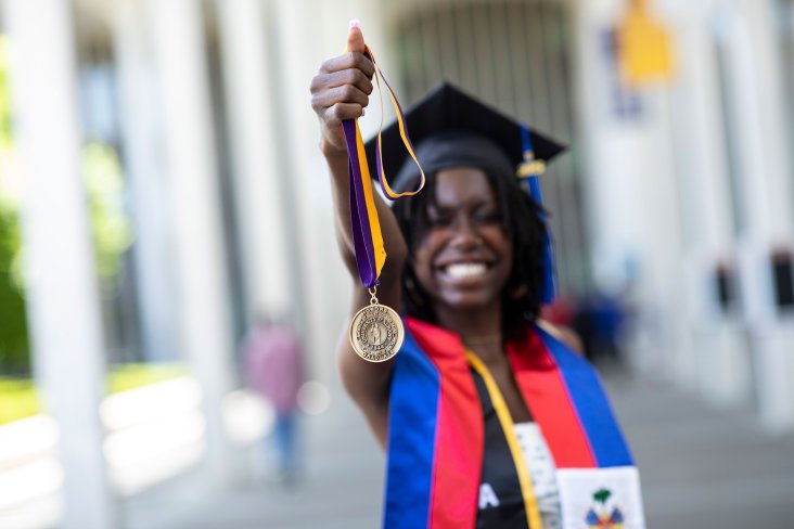 A student wearing a graduate mortar board smiles and holds up an honors medallion.