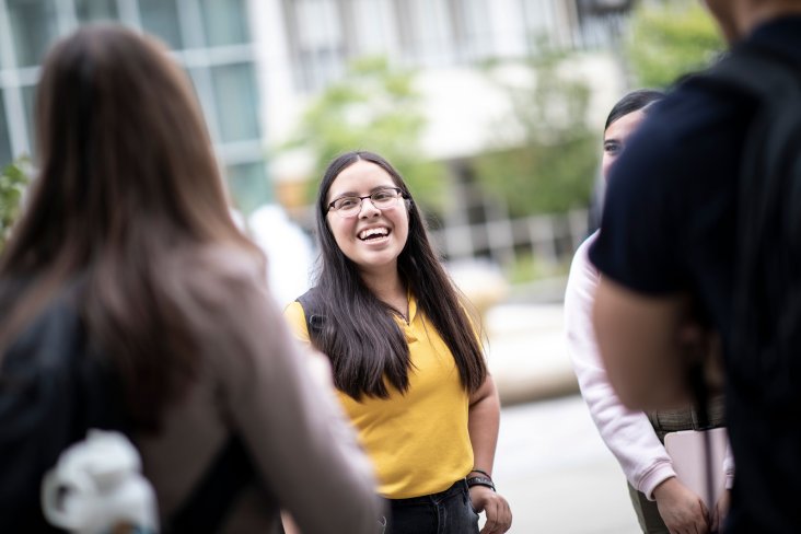 A student in a yellow polo smiles while standing outside with a group of fellow students.