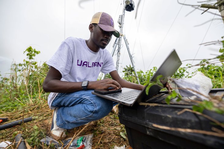 A researcher wearing a UAlbany t-shirt and baseball cap kneels down to work on a computer while installing research equipment in Puerto Rico.