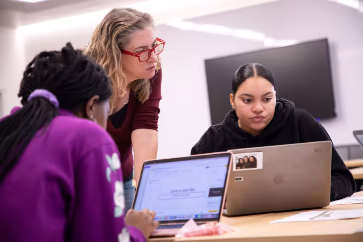 An instructor leans on a table to look at a student's laptop screen. A second student is seated across the table.