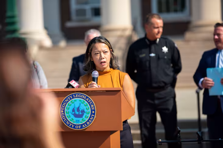 A UAlbany researcher speaks at a podium with the Albany County Legislature seal during an outdoor news conference. County officials stand behind her.