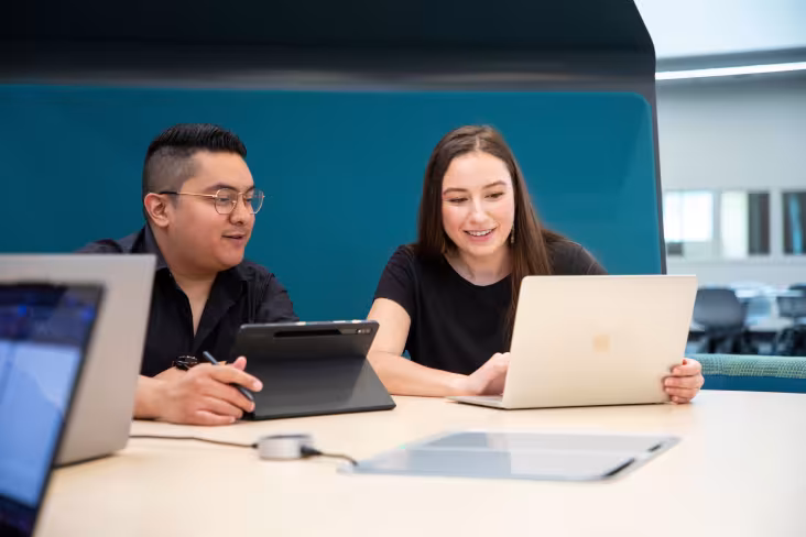Two graduate students smile and chat as they sit at table in front of open laptops.