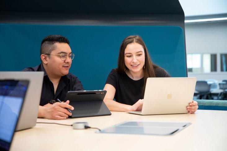 Two graduate students smile and chat as they sit at table in front of open laptops.