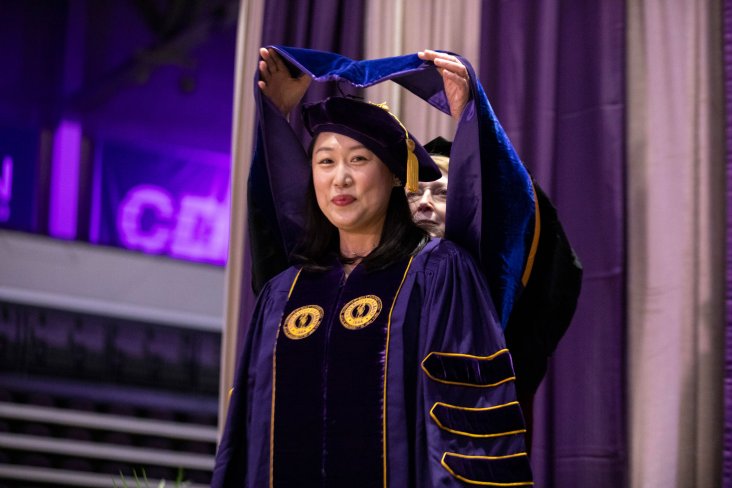 A doctoral student smiles into the camera as a UAlbany academic administrator places a doctoral hood on her shoulders during Commencement.