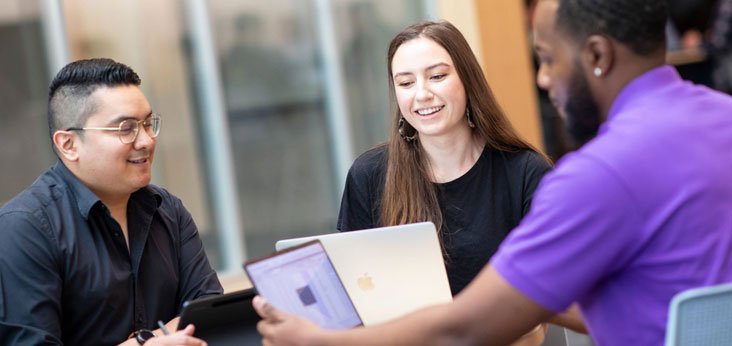 Three smiling people sit around a table in front of laptops.