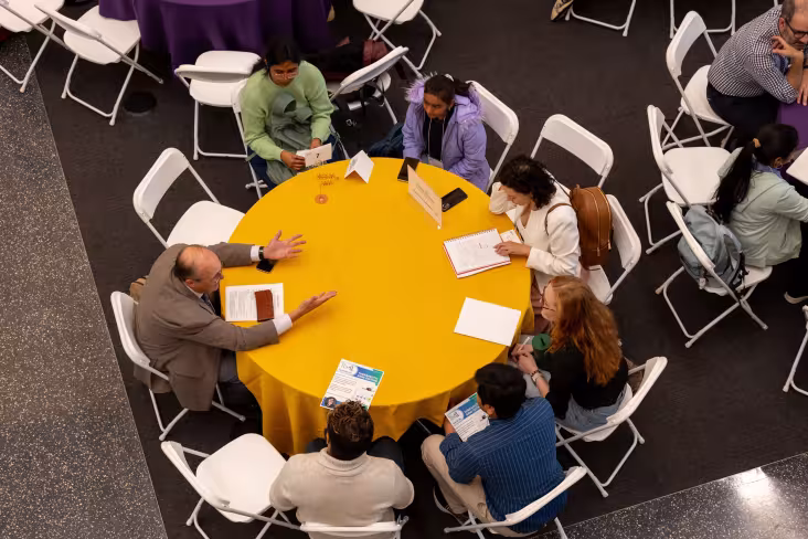 A faculty member and six students sitting at a round table are photographed from above. The researcher gestures as he speaks with the students.