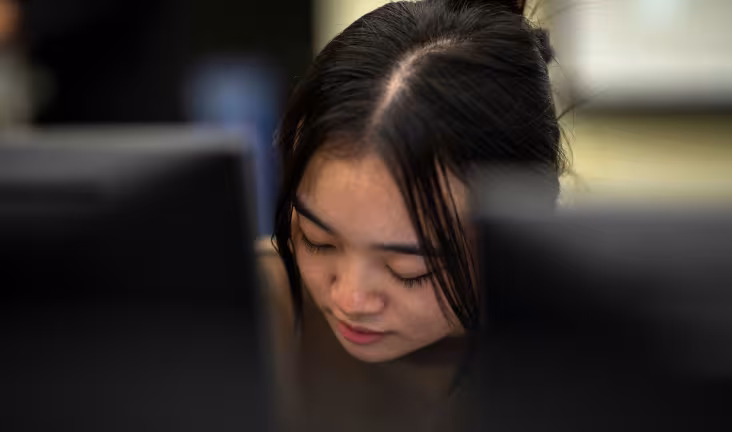 A student sits behind a desktop computer screen, looking down while working.