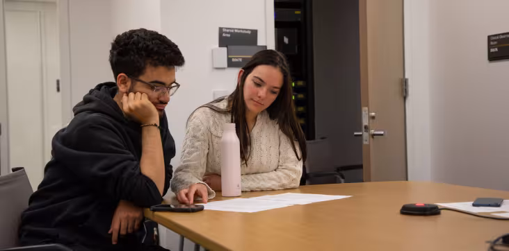 Two students sit at a table inside a tutoring space, looking at paper arrayed before them.