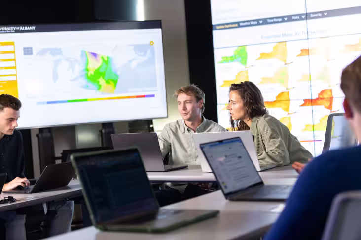 Two students work inside UAlbany's xCITE lab, collaborating on a laptop. Maps of weather patterns in New York State appear on screens behind them.