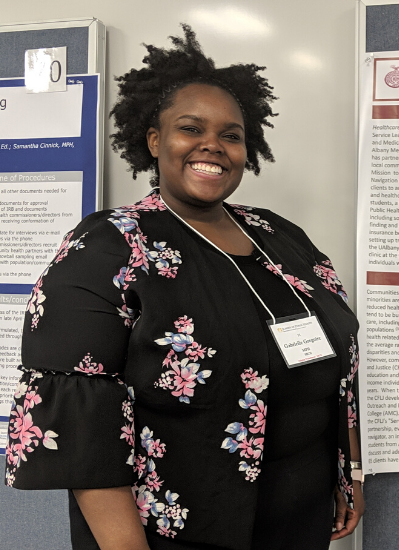 Gabby smiles at the camera, wearing a black top with pink flowers. She is standing in front of her academic poster displaying information from her internship experience.
