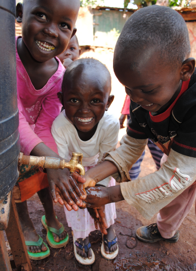 Three children smile while washing their hands outdoors.