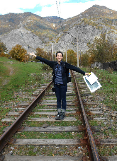 A woman stands on a railroad track in the Country of Georgia, with mountains and grass behind her.