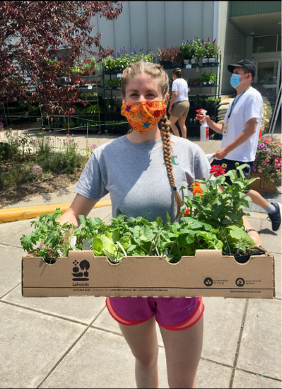 Leanna K. holds a box of planted vegetables. She is wearing an orange face mask and stands outside a childcare center.