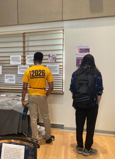 Two people stand in front of a wall displaying project information. The person on the left has a shirt that reads ' Class of 2026", while the person on the right is wearing a blue checked flannel and backpack.