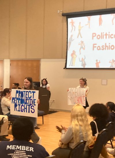 Two women stand in front of a crowd, holding handmade signs reading "Protect Reproductive Rights" and "My Body My Choice". Behind them, "Political Fashion" is displayed on the projector.