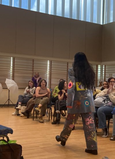 A person with long, black hair walks in front of a crowd of people. She is wearing a painted denim jacked and pants.
