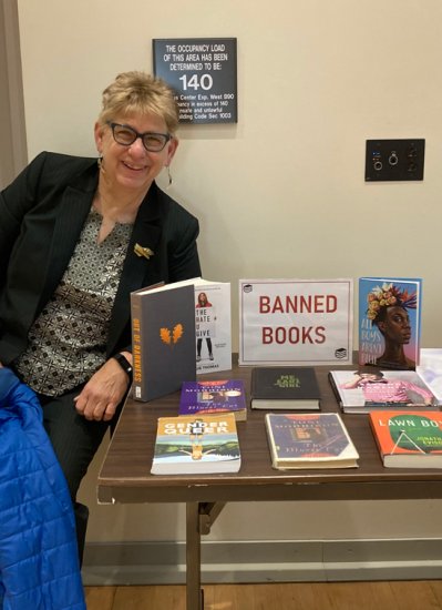 Women in a blazer and glasses poses with a "Banned Books" display table