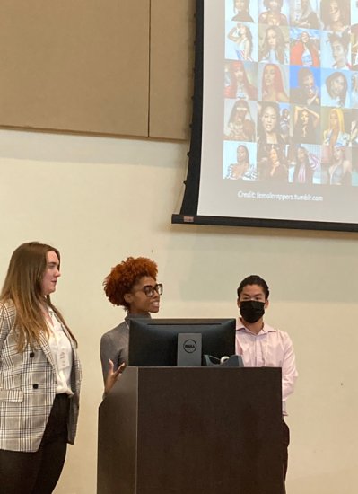 Three people stand behind a podium. The one on the left is in a black and white plaid blazer, the one in the middle is speaking and wearing a gray sweater, and the one on the right is in a pink button-up and a black mask. Behind the, a projector displays a collage of faces with the caption "Credit:femalerappers.tumblr.com"