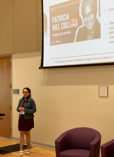 A person in glasses, a denim skirt, and her hair in a ponytail stands in front of a projector with a video queued up. The video that reads thumbnail reads "Patricia Hill Collins" in large font with "Imagens dé controle" underneath.